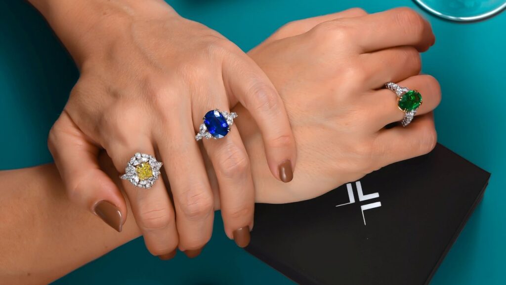 Three custom-made rings by Leibish featuring an oval, 8.51-carat, royal blue sapphire; an oval, 5.49-carat emerald; and a cushion-shaped, 3.74-carat, fancy-green-yellow diamond surrounded by white diamonds.